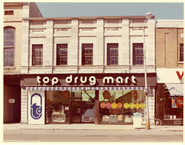 Ein Drogenmart-Gebäude an einer Straßenecke mit einem Schild, einem Straßenschild, einem geparkten Fahrrad, einem Müllcontainer und einem bewölkten Himmel.