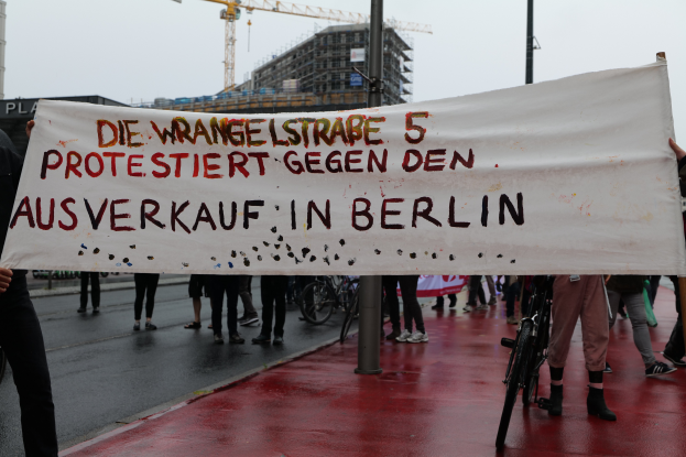 Eine Gruppe von Menschen steht auf der Straße und hält ein Banner mit der Aufschrift "Die Wrangellstraße 5 protestiert gegen den Ausverkauf in Berlin", mit Fahrrädern, Pfählen, Gebäuden und einem Kran im Hintergrund unter einem sichtbaren Himmel.