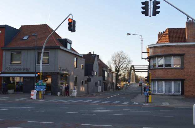 Stadtstraßenkreuzung mit Ampel, umgeben von Gebäuden, Fußgängern, Fahrrädern, Bäumen, Schildern, einem Straßenbogen und einem klaren blauen Himmel.