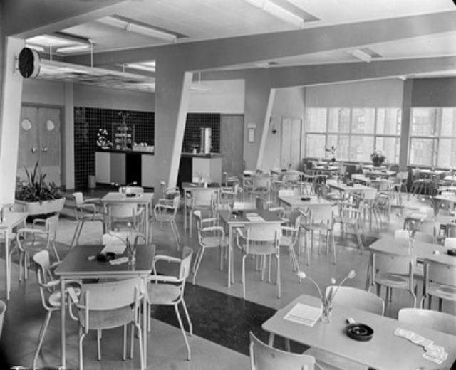 Schwarz-weißes Foto einer Cafeteria mit Tischen und Stühlen, Tellern, Besteck, Gläsern und Blumenvasen auf den Tischen, Türen und Fenstern an der Wand und Deckenleuchten.