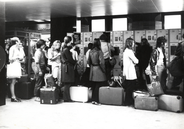 Schwarzes und weißes Bild von Menschen mit Gepäck, die in einem Flughafen stehen, mit Sicht auf Schließfächer im Hintergrund.