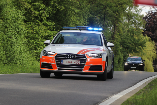 Ein Audi A4 Polizeiwagen fährt auf einer Straße mit Grasrand, flankiert von einem Wald, mit zwei Personen darin, unter einem klaren blauen Himmel.
