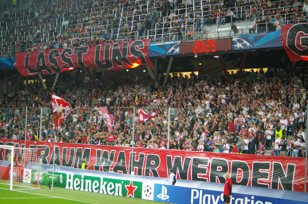 Ein Fuballspiel in einem Stadion mit einer große Menge, saftig grünem Rasen, einem Torpfosten links, Bodenbanner, Menschen mit Fahnen und Hintergrundgeländer, Tafeln, Lichter und andere Gegenstände.