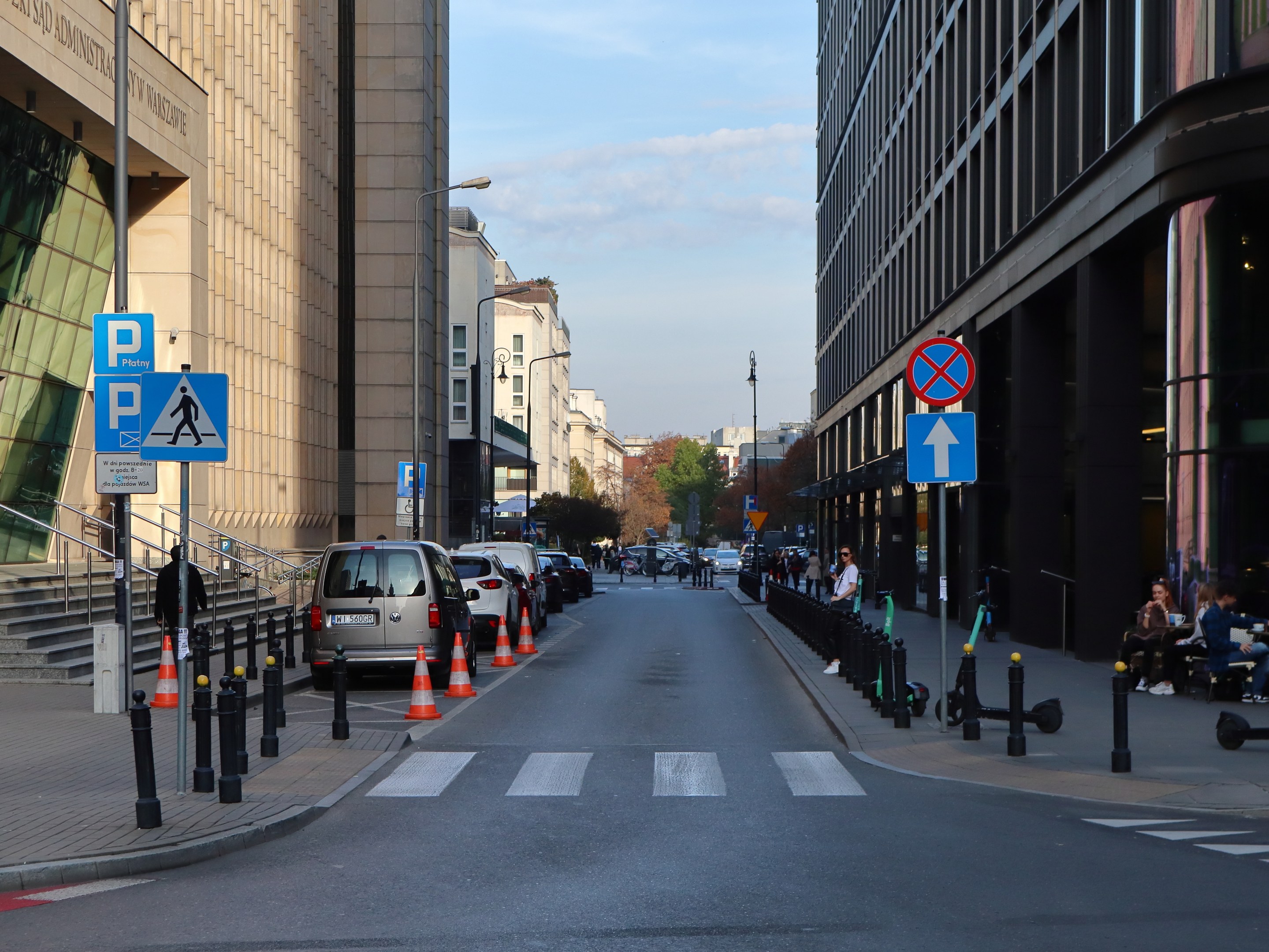 Eine Stadtstraße mit parkenden Autos, Verkehrskegeln, Hinweisschildern, Pfosten, Straßenlaternen, Bäumen und Gebäuden, wo Menschen gehen und auf Bänken sitzen unter einem bewölkten Himmel.
