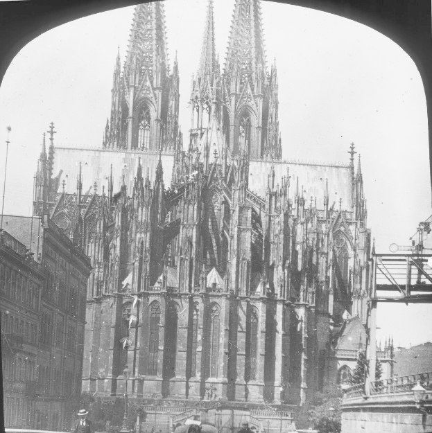 Ein Schwarz-Weiß-Foto des Kölner Doms in Köln, Deutschland, mit seinem hohen Turm, der in den Himmel reicht, umgeben von anderen Gebäuden und Menschen, die auf der Straße vor ihm gehen, mit einer Brücke auf der rechten Seite des Bildes.