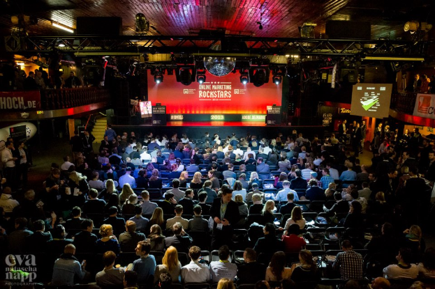 Eine große Menschenmenge sitzt auf Stühlen vor einer Bühne mit Bildschirmen und Bannern, beleuchtet von Scheinwerfern und umgeben von Geländern, mit einem Wasserzeichen in der linken unteren Ecke, das eine Atmosphäre der Aufregung und Vorfreude für ein Online-Marketing-Rockstars-Event schafft.