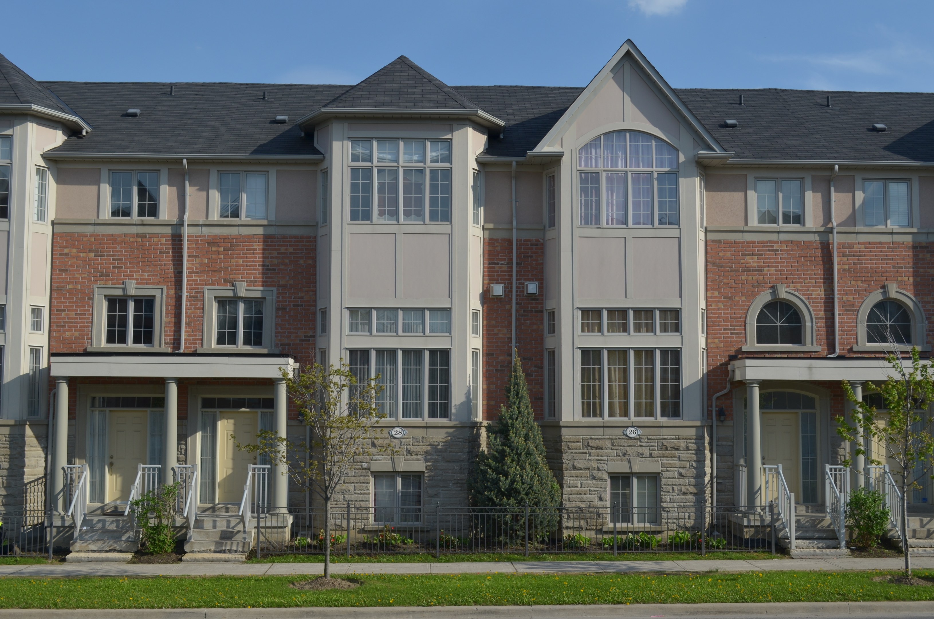 Eine Reihe von Stadthäusern in einem Wohnviertel mit sichtbaren architektonischen Details, Grünflächen und einer Straße im Vordergrund unter einem Himmel als Hintergrund.