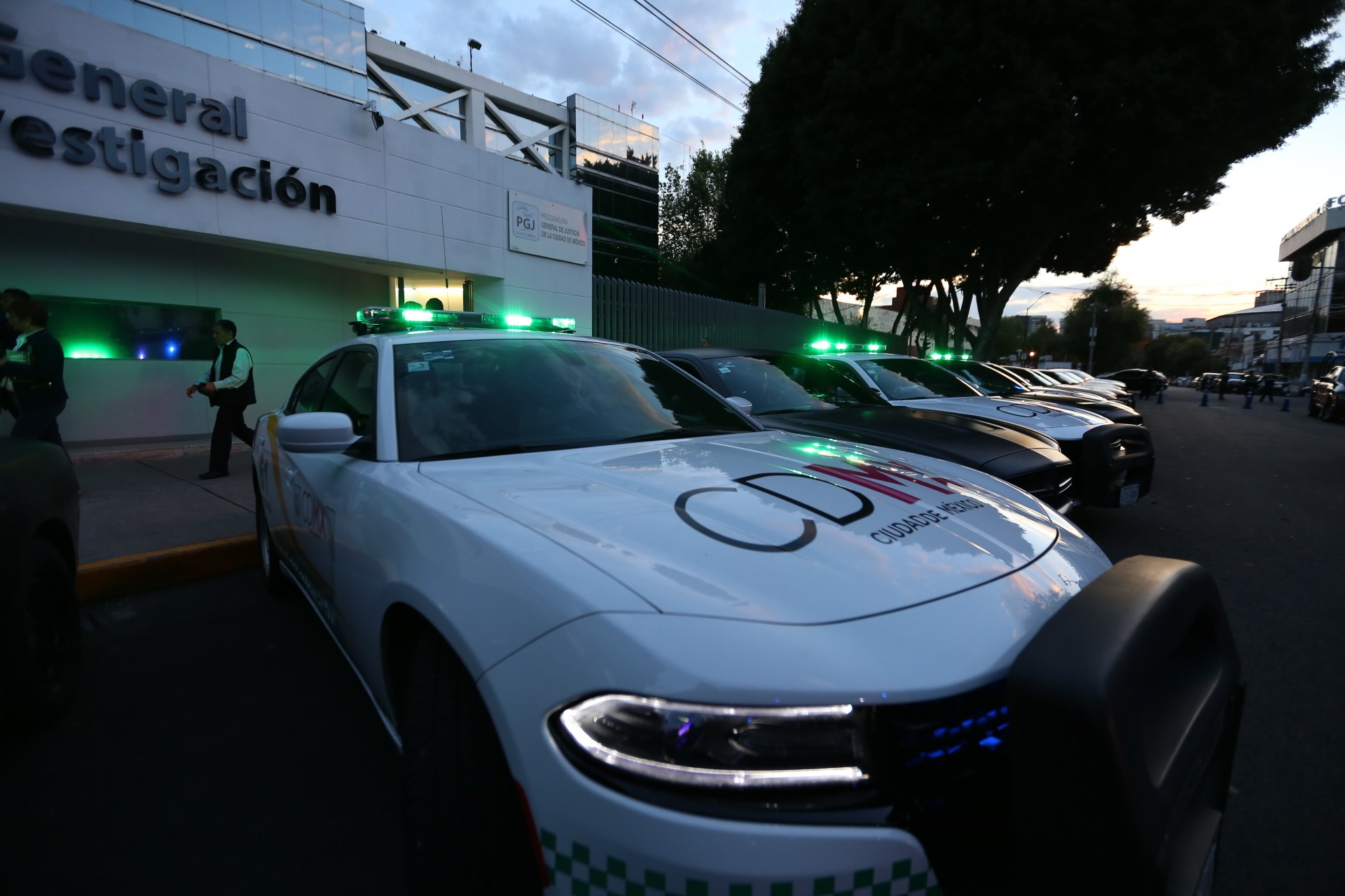 Eine Reihe von Polizeiautos, die vor einem Gebäude mit Text geparkt sind, umgeben von Menschen, Bäumen, Laternenmasten, Drähten und einem bewölkten Himmel.
