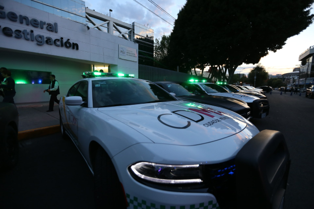 Eine Reihe von Polizeiautos, die vor einem Gebäude mit Text geparkt sind, umgeben von Menschen, Bäumen, Laternenmasten, Drähten und einem bewölkten Himmel.