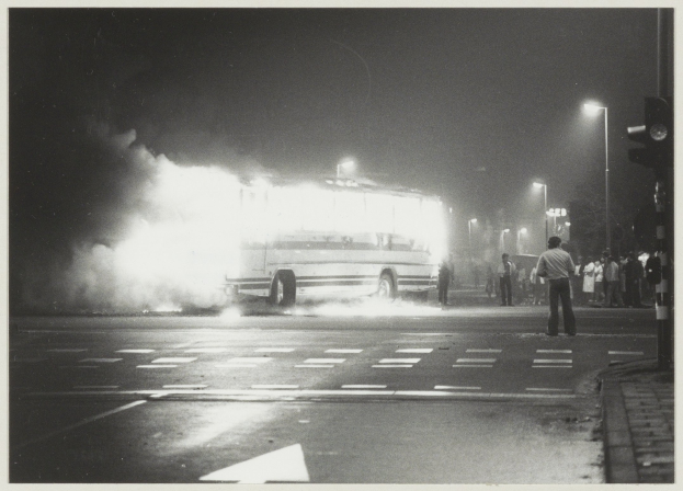 Ein Schwarz-Weiß-Foto eines brennenden Busses am Straßenrand, mit einer Gruppe von Menschen auf dem Gehweg rechts daneben und Straßenlaternen im Hintergrund.
