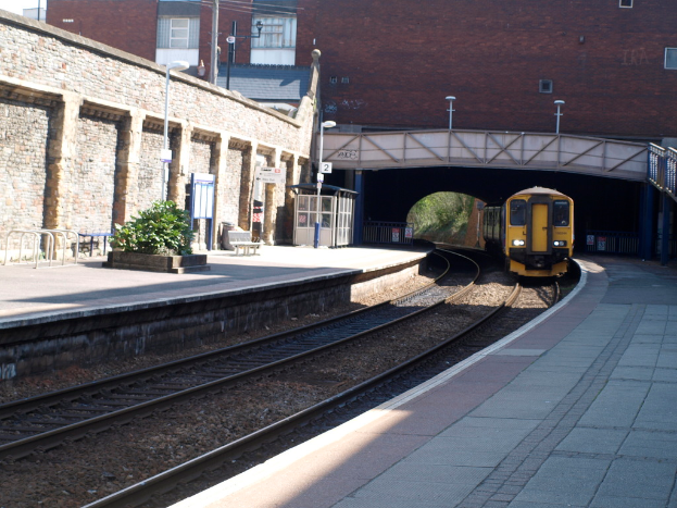 Ein Zug fährt auf den Schienen mit Plattformen auf beiden Seiten, eine Hauswand links, Straßenlaternenpfosten, Pflanzen, Bänke, eine Kabine, eine Brücke über die Plattformen und Gebäude und Pfosten im Hintergrund.