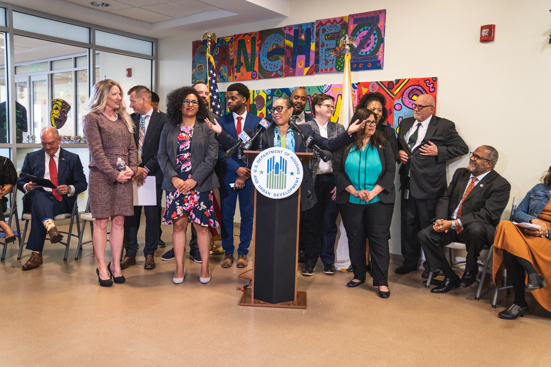 Gruppe von Menschen vor einem Podium mit Mikrofon und einer Tafel mit der Aufschrift 'Sancho-Stadtrat tritt dem Stadtrat bei', mit sitzenden und stehenden Teilnehmern, Fahnen, Feuerlöscher und Deckenbeleuchtung im Hintergrund.