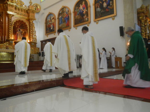 Eine Gruppe von Priestern geht einen roten Teppich in einer Kirche entlang, mit Menschen auf dem Boden und auf der rechten Seite, und Fotorahmen im Hintergrund.