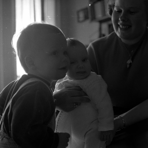 Eine Frau hält ein Baby mit einem warmen Lächeln, mit zwei anderen Kindern daneben, in einem gemütlichen Raum mit Rahmen und einem Fenster im Hintergrund.