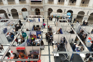 Eine große Gruppe von Menschen versammelt sich in einem Raum mit Ständen, Bannern, Tischen, Stühlen, Luftballons und einem Gebäude im Hintergrund.