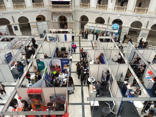 Eine große Gruppe von Menschen versammelt sich in einem Raum mit Ständen, Bannern, Tischen, Stühlen, Luftballons und einem Gebäude im Hintergrund.