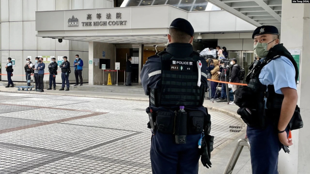 Zwei Polizisten in Uniform, Mützen und Masken stehen vor einem Gebäude am Hong Kong International Airport, umgeben von einer Gruppe von Menschen, einige halten Kameras, mit Stangen, die mit Bändern geschmückt sind, im Hintergrund.