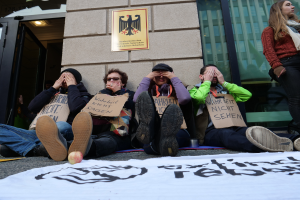 Eine Gruppe von Menschen sitzt vor einem Gebäude auf dem Boden, hält Schilder, mit einer Fahne und einem Apfel auf der Straße, und eine Frau steht rechts daneben, sieht besorgt aus.