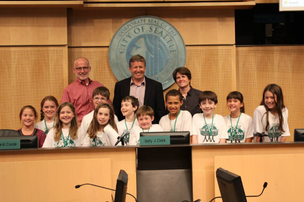 Eine Gruppe von Kindern und Erwachsenen steht vor einem Rednerpult, lächelt und posiert für ein Foto, mit Monitoren, Mikrofonen und anderen Gegenständen auf einem Tisch im Vordergrund, einem Logo an der Wand und einem Bildschirm auf der rechten Seite im Hintergrund, bei einer Stadtratssitzung.