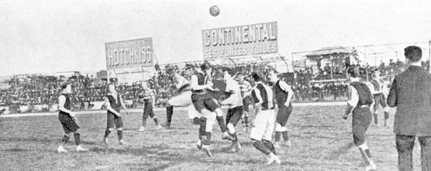 Ein Schwarz-Weiß-Bild einer Gruppe von Männern, die auf einem Feld Fußball spielen, mit einem Ball in der Luft und Zuschauern im Hintergrund, mit Tafeln, die Text anzeigen.