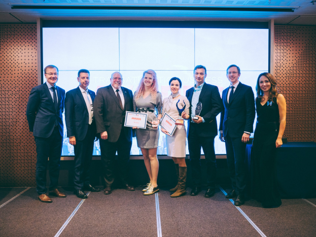 Gruppe strahlender Menschen mit Urkunden, vor einem Bildschirm mit einem Tisch und einem wandmontierten Objekt im Hintergrund, Gewinner der Internationalen Wirtschaftsauszeichnungen 2018.