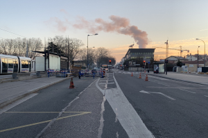 Eine Stadtstraße mit Verkehrskegeln, einem Bus, Laternenmasten, Bäumen, Gebäuden, Kränen und Rauch, der aus einer fernen Struktur aufsteigt.