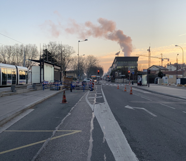 Eine Stadtstraße mit Verkehrskegeln, einem Bus, Laternenmasten, Bäumen, Gebäuden, Kränen und Rauch, der aus einer fernen Struktur aufsteigt.