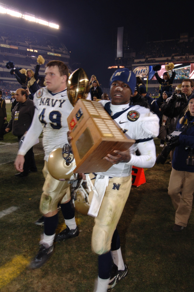 Ein Fußballer mit einem Pokal auf einem Feld, umgeben von einer Menge in Sportkleidung, mit Schildern, Lichtern und einem klaren blauen Himmel im Hintergrund.