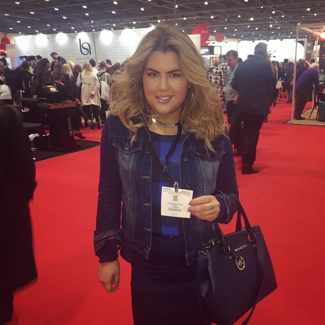 Eine Frau in einem roten Kleid steht auf einem roten Teppich, lächelt und hält eine Handtasche, während im Hintergrund Menschen, Lichter und Schilder zu sehen sind.