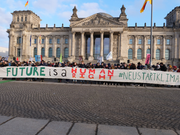 Gruppe von Menschen mit einem Banner "Zukunft ist ein Mensch" vor dem Reichstaggebäude in Berlin, Deutschland, mit seinen architektonischen Details und umgeben von Fahnen unter einem bewölkten Himmel.