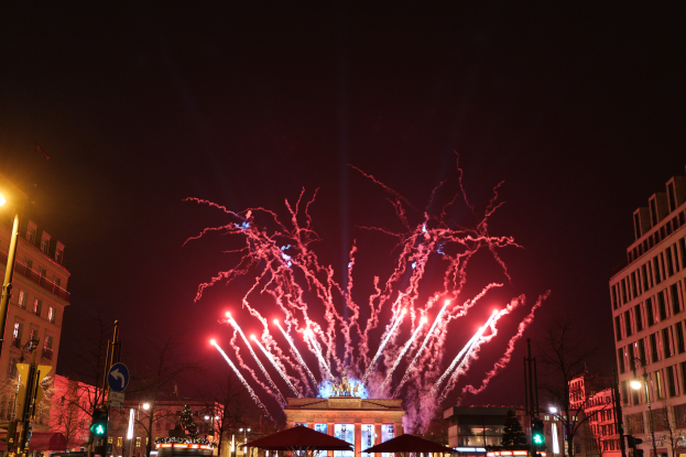 Eine belebte Stadtstraße in Berlin an Silvester, voller Menschen, Fahrzeuge und Gebäude, erhellt von Feuerwerk und Gebäudebeleuchtung, die eine festliche Stimmung schaffen.