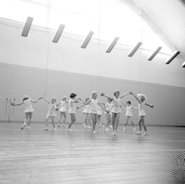 Schwarz-weißes Bild von jungen Mädchen, die sich an den Händen halten und in einem Studio tanzen, mit einem Ball auf der rechten Seite, Deckenbeleuchtung und einem Wandobjekt im Hintergrund.