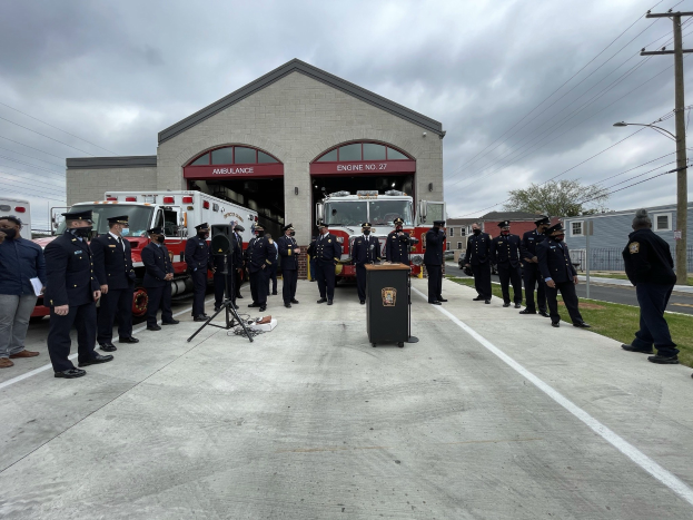 Gruppe von Menschen in Uniformen neben einem Feuerwehrauto und einem Rednerpult bei einer Zeremonie für eine neue Feuerwache, mit Gebäuden, Bäumen, Strommasten, Wolkenhimmel im Hintergrund.