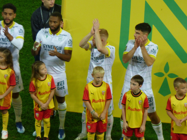 Eine Gruppe junger Menschen in Fußballuniformen steht auf einem Fußballfeld und klatscht feierlich mit einer 'Ligue 1'-Tafel im Hintergrund.