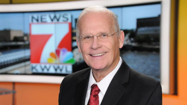 Ein Mann im Anzug und Krawatte, der eine Brille trägt, steht vor einem Fernseher und lächelt, mit einem unscharfen Hintergrund.