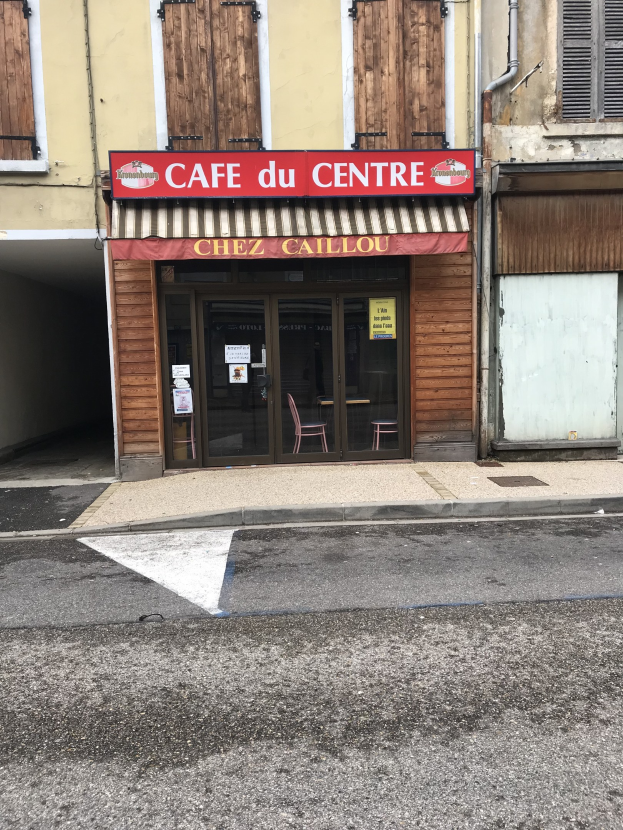 Ein zweistöckiges Gebäude mit einer 'Cafe du Centre'-Schriftzug an der Seite, das Fenster, eine Tür und ein kleines Straßenbrett mit Text davor zeigt.
