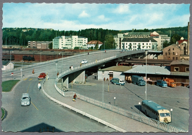 Altes Schwarz-Weiß-Foto einer Stadtstraße mit Fahrzeugen, Fußgängern auf einer Brücke, Gebäuden, Bäumen, Laternenpfählen und einem bewölkten Himmel.