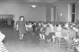 Eine Frau in einem schwarzen und weißen Kleid läuft über den Laufsteg während einer Modenschau für Frauenwahlrecht, mit einer Menge, die auf Stühlen sitzt und einer Uhr und Lichtern an den umliegenden Wänden.