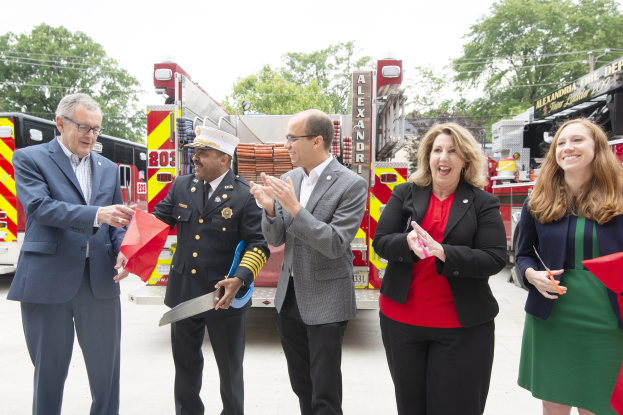 Group of people at a ribbon cutting ceremony for the Alexandria Fire Department, clapping and smiling in front of a fire truck, with two individuals holding scissors and a red ribbon.