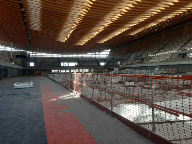 Großes Stadioninnere im Bau mit sichtbarem Boden, Deckenbeleuchtung und einem Netzzaun auf der rechten Seite; Hintergrund zeigt wenige Details.