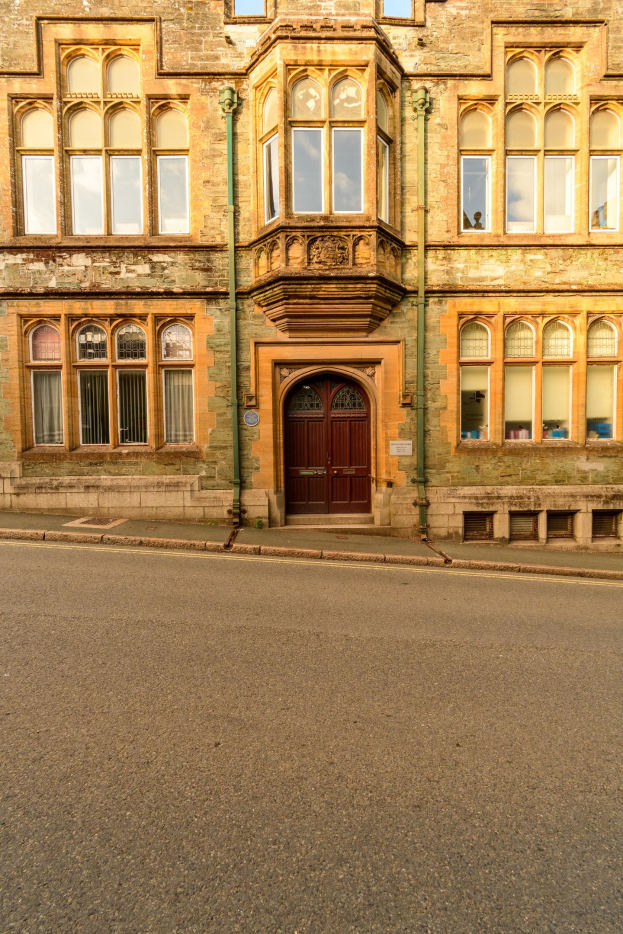 Alter Bau mit vielen Fenstern und einer hölzernen Tür, neben einer Straße gelegen und mit Rohren an seinen Wänden.