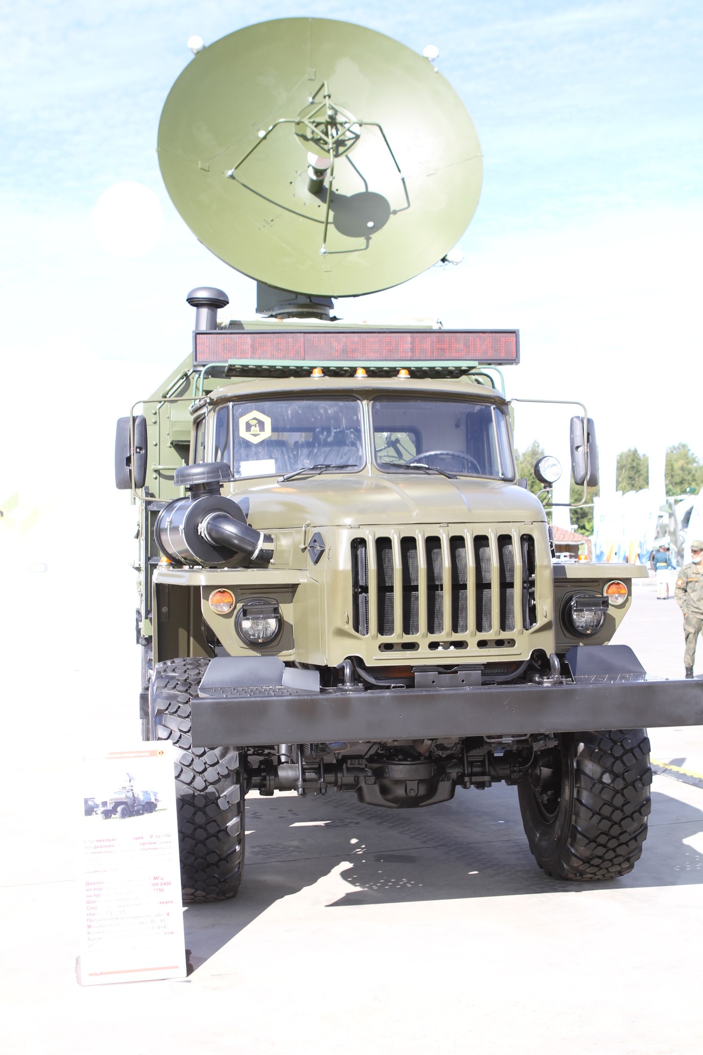 Militärlastwagen mit einer montierten Satellitenschüssel, auf einem Parkplatz mit Menschen, Bäumen und einem klaren blauen Himmel im Hintergrund.