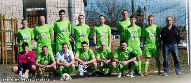 Gruppe von Männern in grünen T-Shirts, die auf einem Fußballfeld stehen oder kauern, mit einem Ball und einem Torpfosten umgeben von Bäumen, Gebäuden und Fahrzeugen.