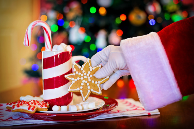Mann in einem Santa Claus Hut, der eine Tasse heißer Schokolade und einen Zuckerstange hält und einen Teller mit Plätzchen und ein Tuch auf dem Tisch vor sich hat, vor einem Weihnachtsbaum und einer Wand.