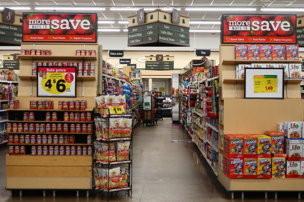 Ein Kiosk-Innenraum mit Regalen und Bodenbestand an verschiedenen Lebensmitteln, Deckenbeleuchtung und einer Person im Hintergrund.