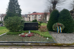 Ein Denkmal in einem Park mit einem Kranz, umgeben von Gras, Pflanzen, Blumen und Bäumen, mit Gebäuden und dem Himmel im Hintergrund, gewidmet den Opfern des Zweiten Weltkriegs.