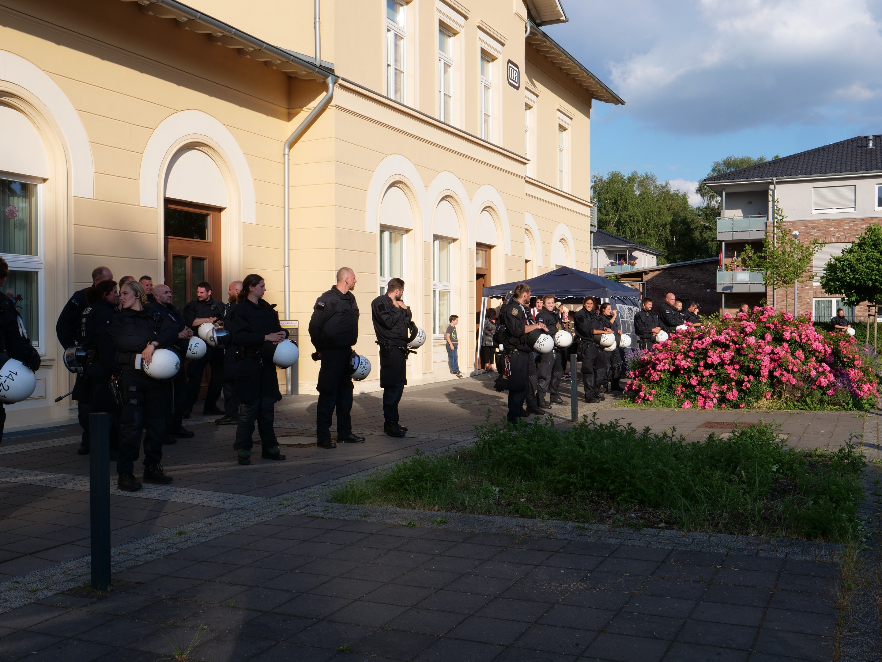 Gruppe von Polizisten vor einem Gebäude mit Fenstern und Türen stehend, Helme haltend, in der Nähe eines Zeltes, mit Grünfläche, einer Straßenlaterne, Bäumen und einem bewölkten Himmel.