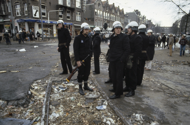Eine Gruppe von Polizisten in Helmen und mit Waffen in den Händen steht an der Seite einer Straße mit Gebäuden, Schildern, Laternen, Bäumen und einem klaren Himmel im Hintergrund.