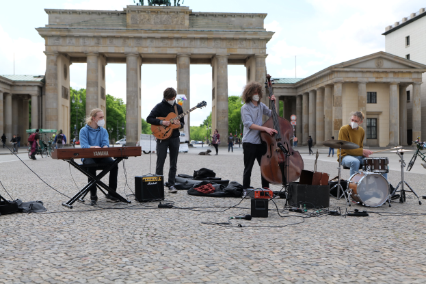 Gruppe von Musikern, die draußen spielen, mit einem Gitarristen im Sitzen, einem anderen Musiker, der ein Instrument spielt, und einer Person mit Maske vor dem Brandenburger Tor mit verstreuter Ausrüstung und Zuschauern im Hintergrund.