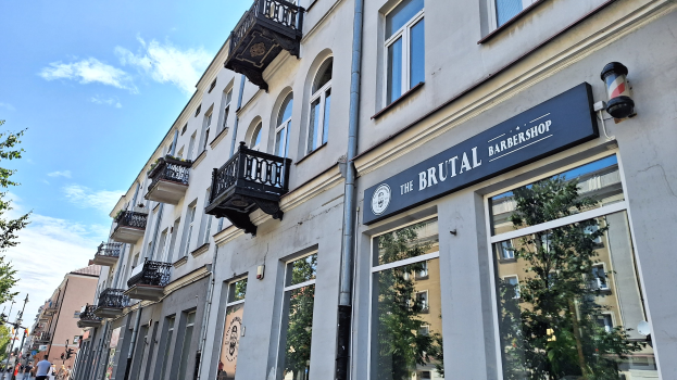 Brutalistischer Barbershop-Außenbereich in Berlin mit eckigen Betongebäuden, Fenstern, Geländern, Balkonen und Namensschildern, umgeben von Bäumen, Laternenmasten und Fußgängern an einem bewölkten Tag.